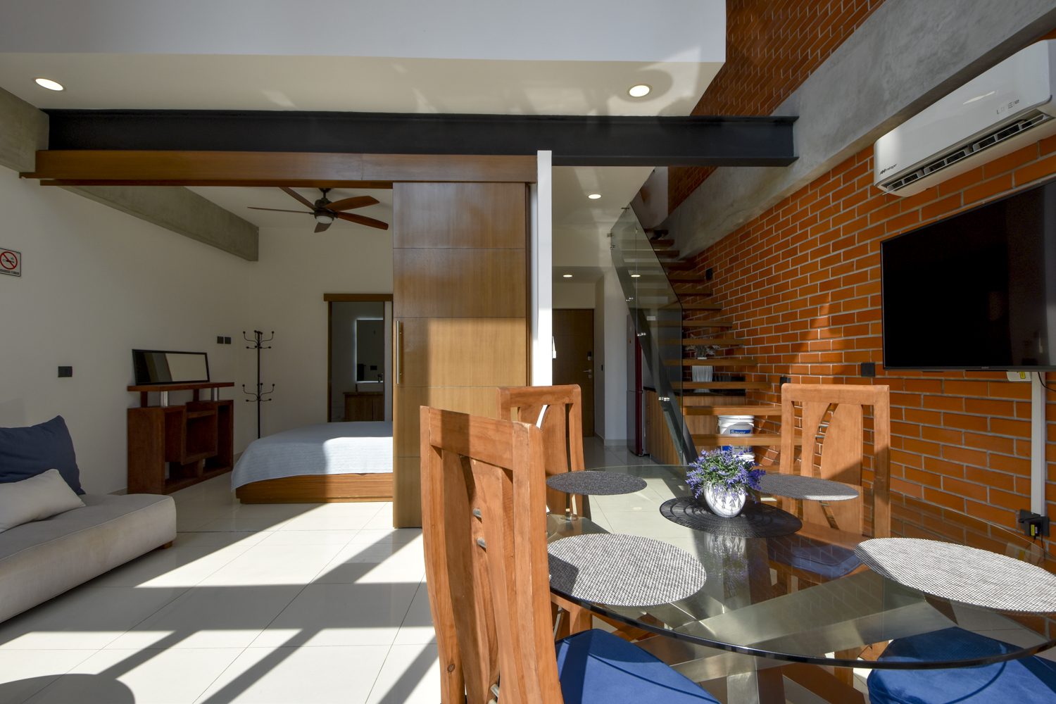 Open loft living area of Zoho Skies 108 with exposed brick wall, dining table, sofa, bedroom area and floating staircase